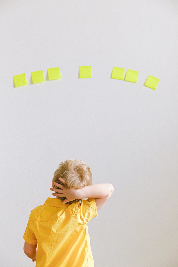 A young boy in a yellow shirt ponders yellow sticky notes on a white wall.